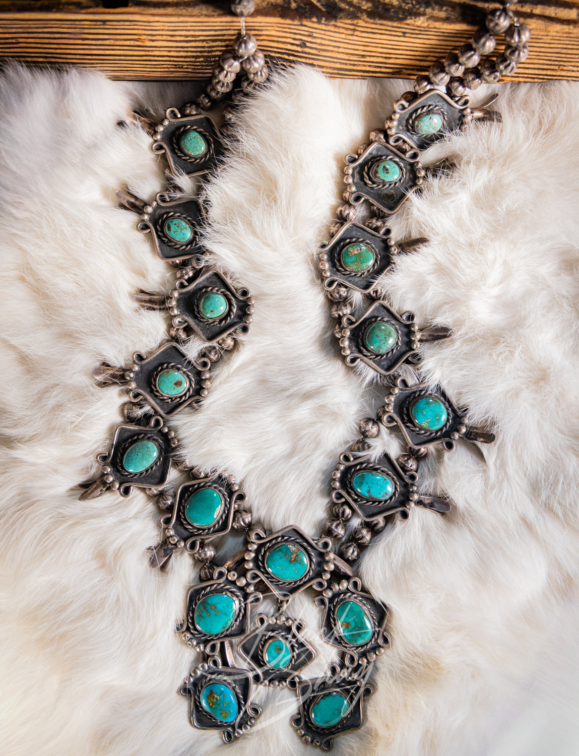 A Southwestern-style silver-toned necklace with multiple turquoise stones, displayed on a white faux fur surface with a wooden board at the top.