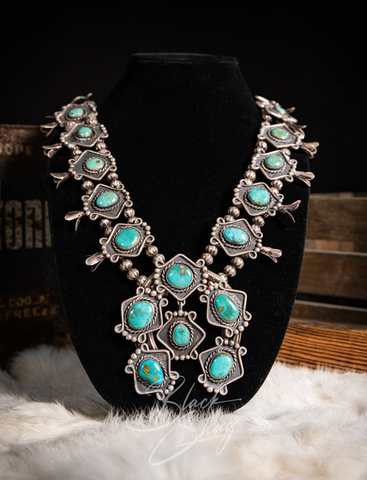 A silver-toned Southwestern-style necklace with multiple turquoise stones, displayed on a black velvet jewelry bust. The necklace features intricate silver bezel settings and is styled on a white faux fur surface with rustic wooden boxes in the background.