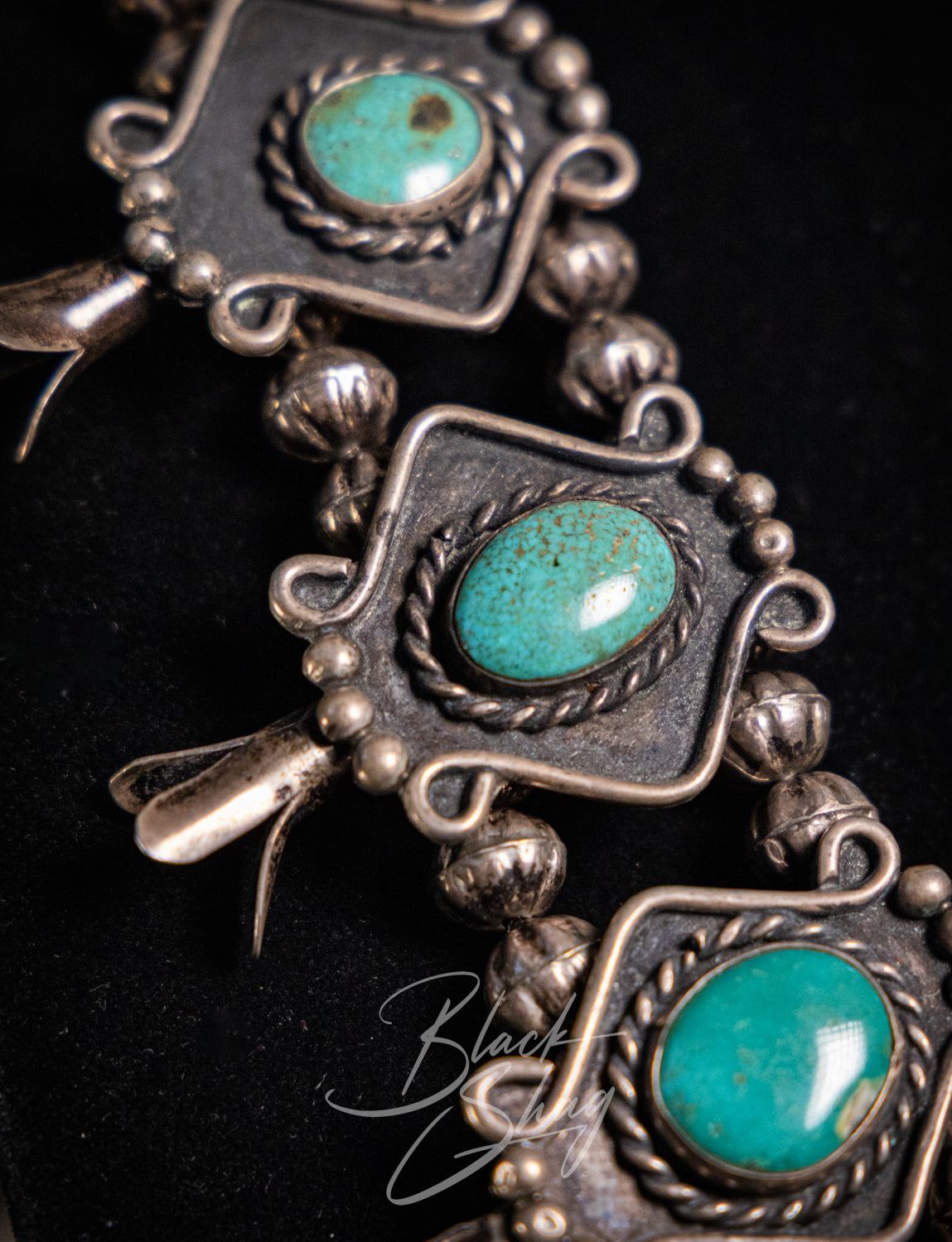 A close-up of a silver-toned jewelry piece featuring three oval turquoise stones set in ornate, carved metal frames with decorative beads and dangling elements, photographed against a black background.