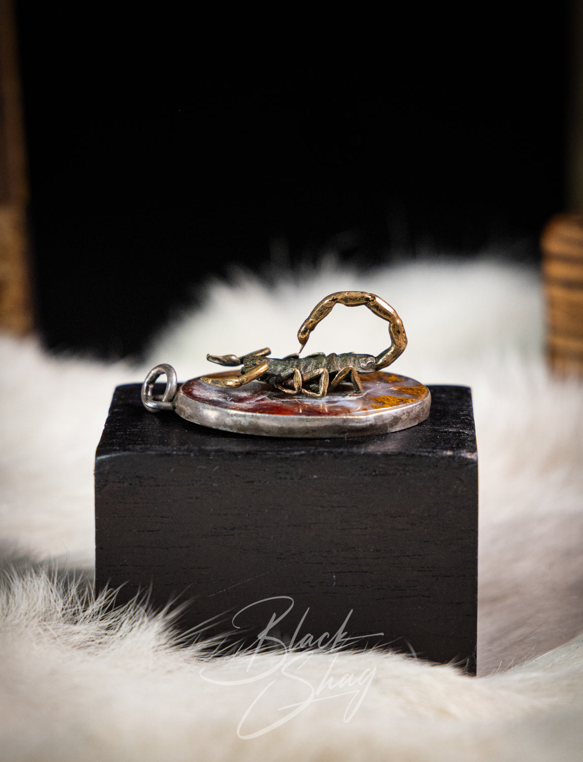 A silver pendant featuring a detailed gold-toned scorpion figurine standing on a circular amber-colored background, displayed on a black block over a white faux fur surface.