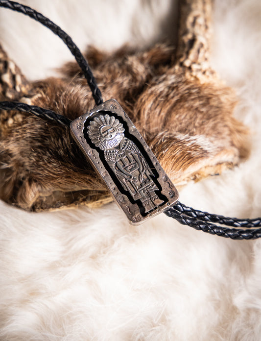 A close-up of a braided black cord necklace with a rectangular engraved metal pendant featuring a stylized figure, styled over brown fur on a white faux-fur surface.