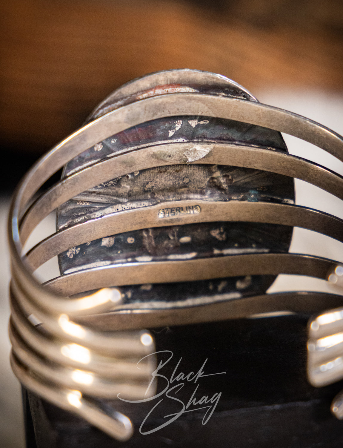 A close-up of a silver-toned metal cuff bracelet with multiple parallel bands and a textured dark inlay, photographed against a blurred warm brown background. A script watermark reading 'Black Shag' is visible near the bottom.