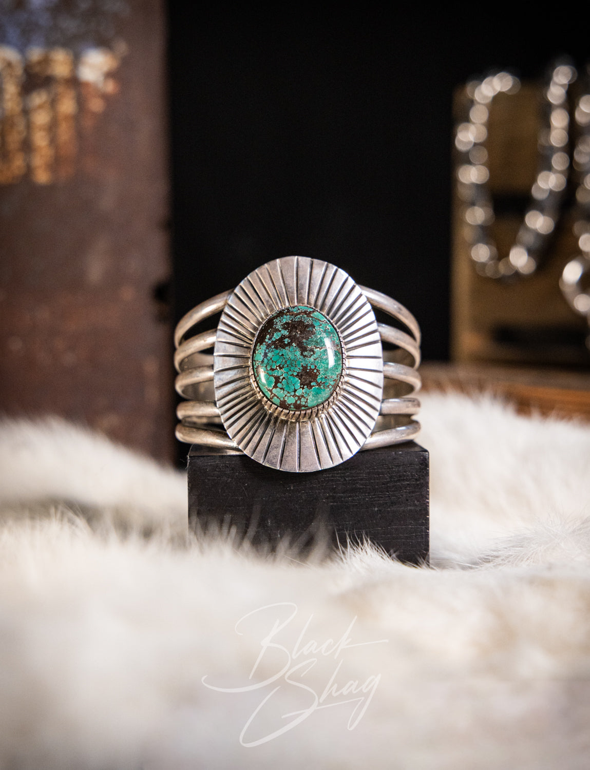 A sterling silver cuff bracelet with a round turquoise stone centerpiece, displayed on a black block over a white faux fur surface.