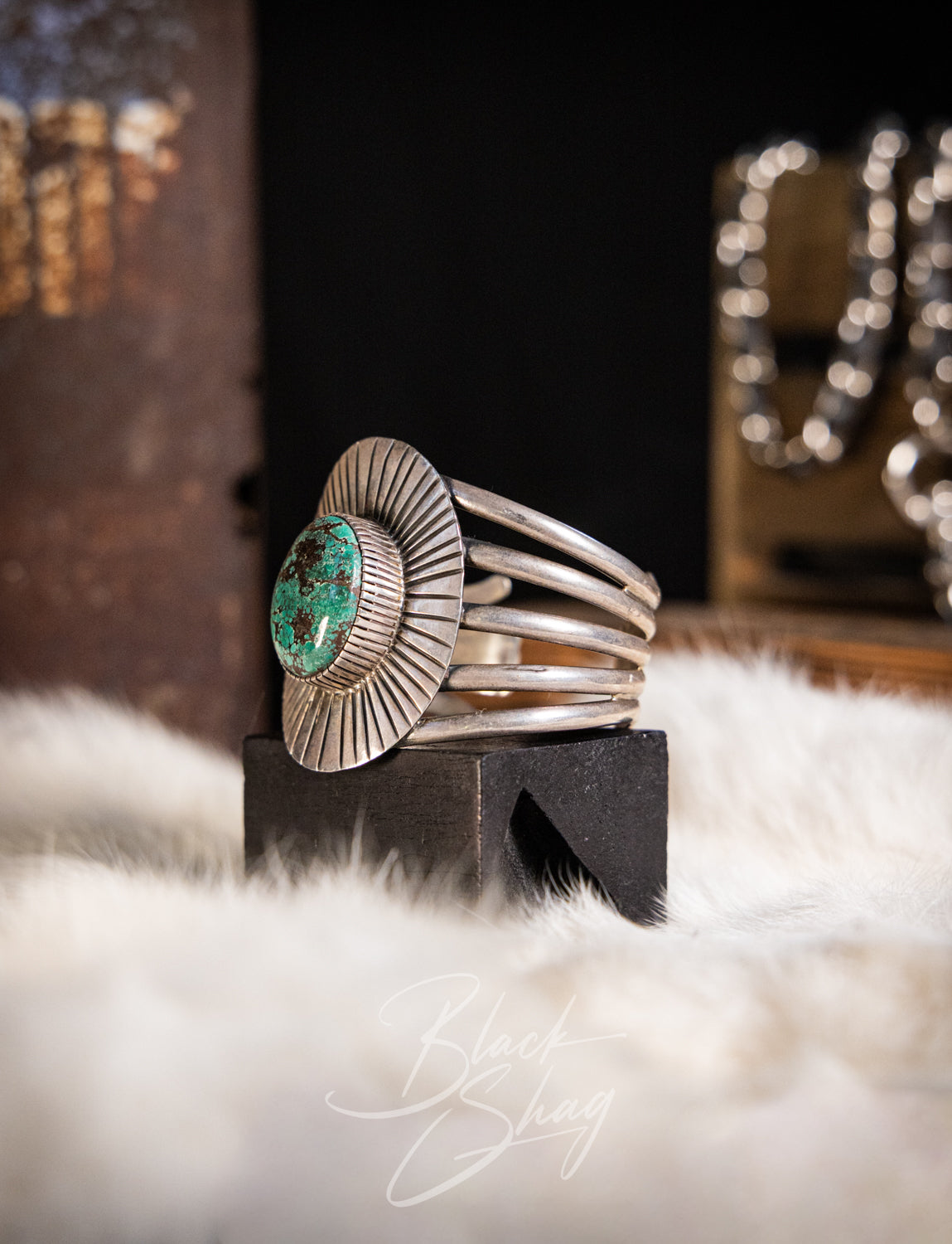 A sterling silver band ring with a textured sunburst face and a central green turquoise stone, displayed on a black block over a white faux fur surface.