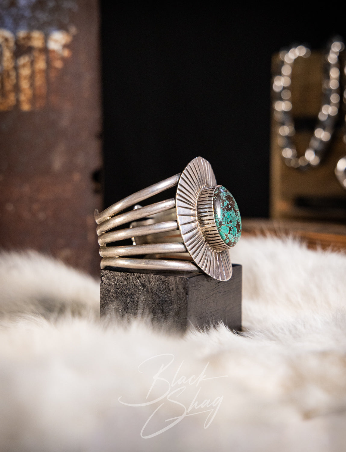 A wide silver cuff ring with a round turquoise stone set in a sunburst bezel, displayed on a black block over a white faux fur surface.