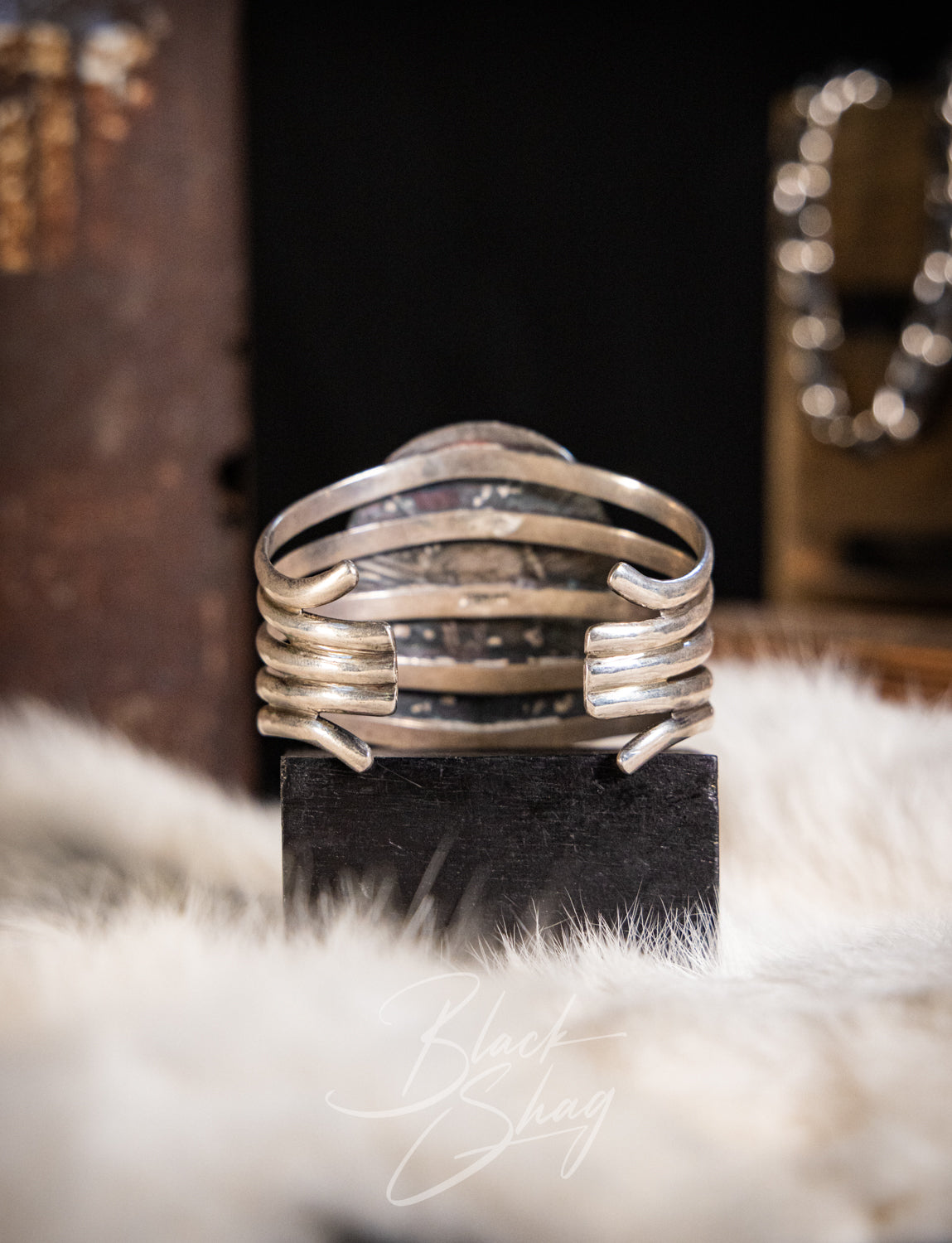 A wide silver cuff ring with multiple horizontal bands and a textured center, displayed on a black block over a white faux fur surface.