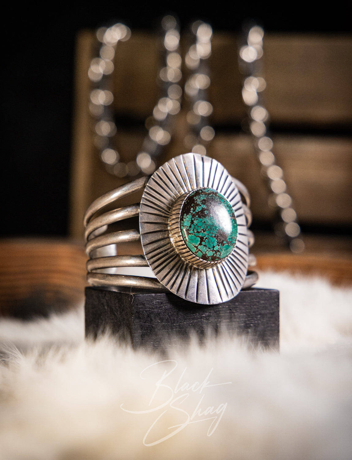 A sterling silver cuff ring with a large oval green turquoise stone, displayed on a black block over a white faux fur surface with a blurred wooden background.
