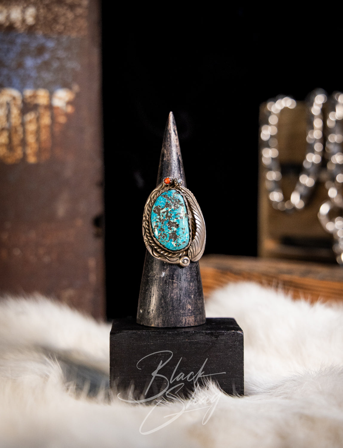 A silver-toned ring with a turquoise-blue oval stone and small orange accent, displayed on a black cone stand with a dark background and white faux-fur surface.