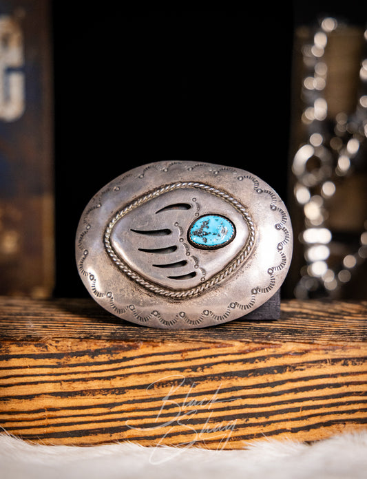 A silver-toned Southwestern-style concho buckle with a turquoise stone inlay, displayed on a rustic wooden surface with a dark background.
