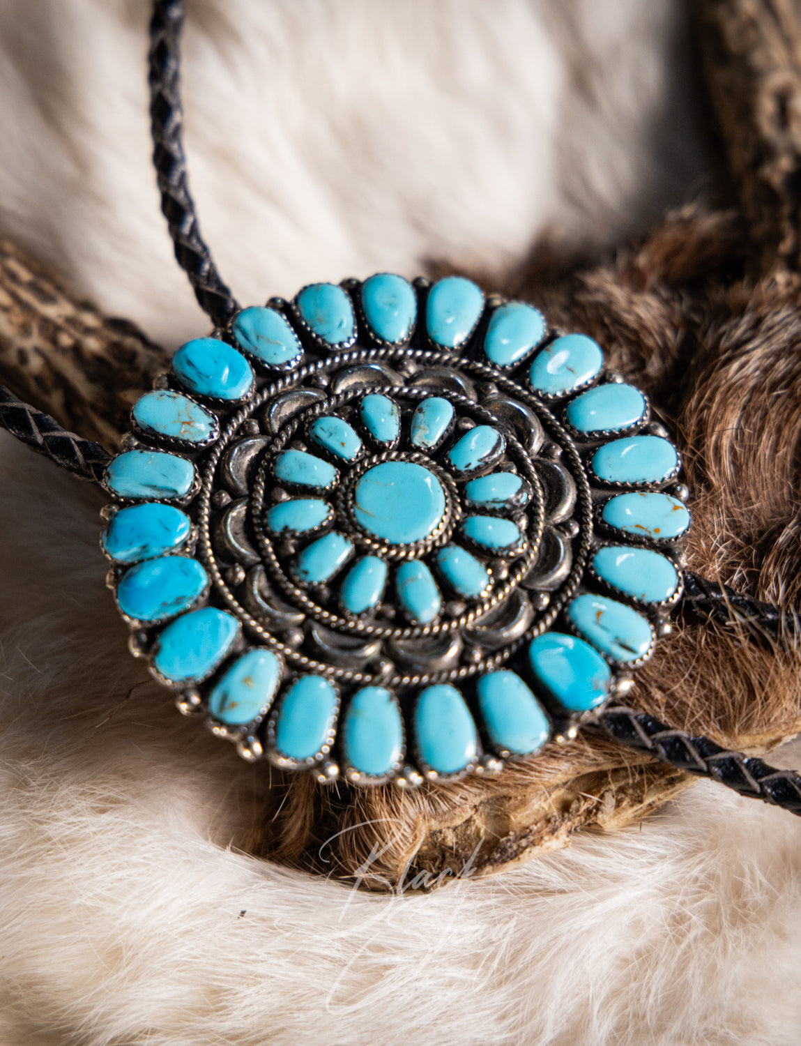 A close-up of a circular turquoise and silver-toned bolo-style necklace laid on a soft white and brown fur surface.