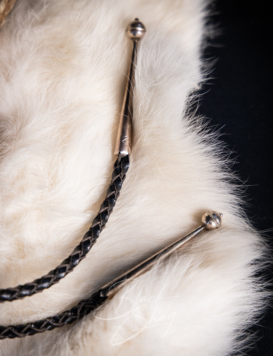 Close-up of a black braided strap with silver-toned metal ends and a small round bead, resting on white fur against a black background.