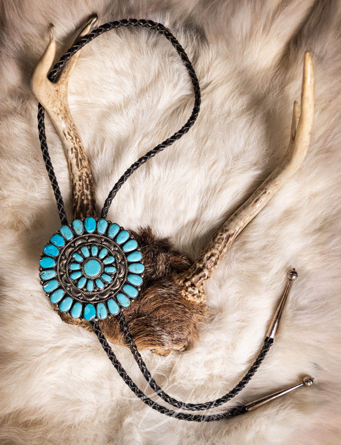 A rustic Native American style bolo tie featuring a circular turquoise and bronze inlay centerpiece, suspended on a black braided cord with antler and fur elements, laid flat on a white fur background.