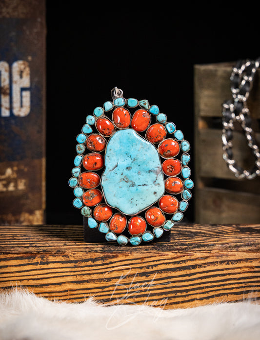A Southwestern-style turquoise and coral pendant necklace displayed on a wooden stand. The pendant features a large, irregular light blue turquoise center stone surrounded by a border of small turquoise and orange-red coral stones set in silver-tone metal.