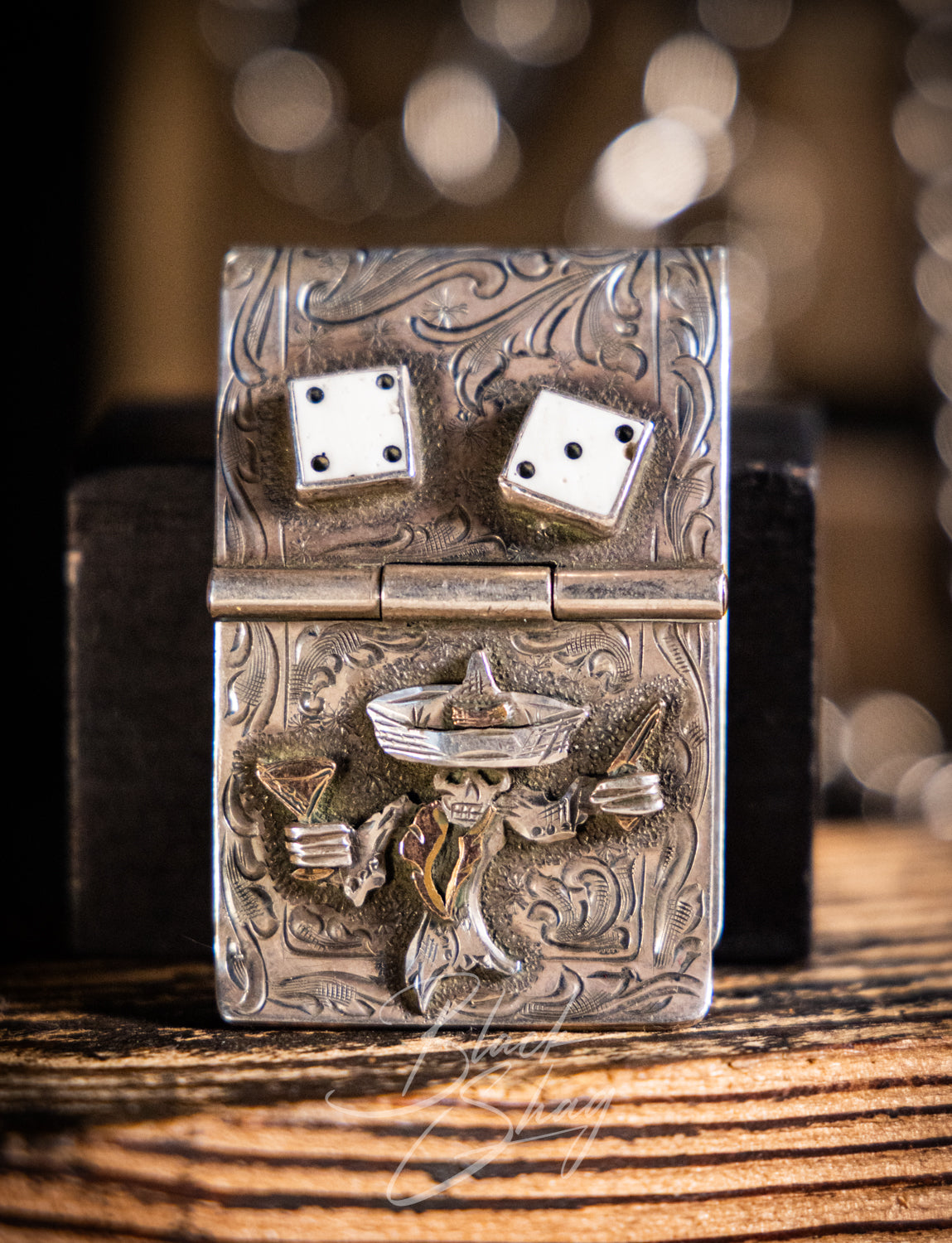 A silver-toned engraved lighter with a Mexican sombrero and dice design, resting on a wooden surface with a dark box behind it.