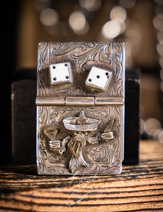 A silver-toned engraved lighter with a Mexican sombrero and dice design, resting on a wooden surface with a dark box behind it.