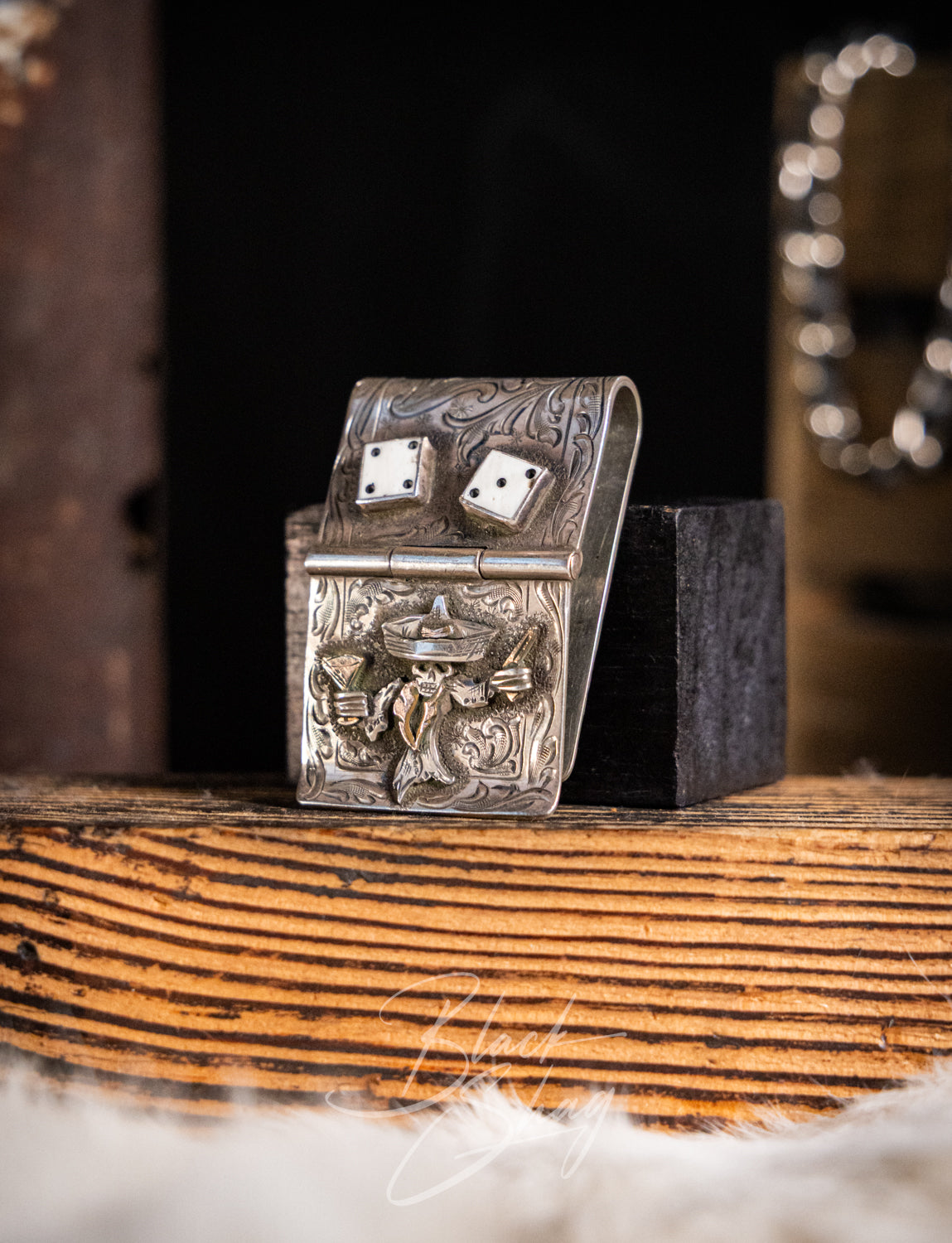 An ornate silver-toned metal dice box with engraved decorative artwork and two visible dice, displayed on a rustic wooden surface with a dark block behind it.
